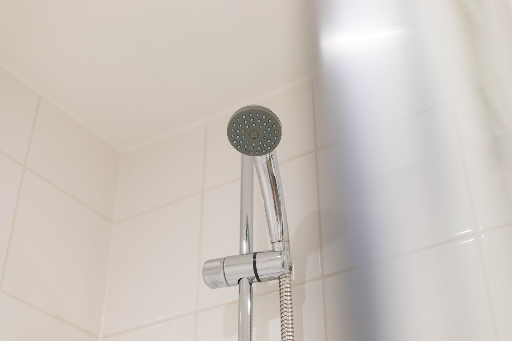 Un pommeau de douche doté d’un flexible, fixé sur un accessoire chromé, dans une salle de bain aux murs recouverts de carreaux blancs.