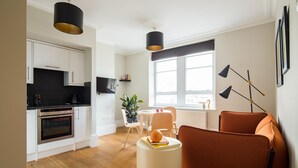 A modern kitchen with white cabinets, a black countertop, and stainless steel appliances. There is a dining area with a table, chairs, and a potted plant. The room has a large window and a floor lamp. - Cool Covent Garden 1BR - Netflix & Nespresso (London)