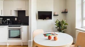 A modern kitchen with white cabinets, a black countertop, and a built-in oven. There is a round table with two apples and a magazine, and a potted plant. - Cool Covent Garden 1BR - Netflix & Nespresso (London)