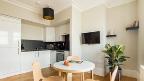 A modern kitchen with white cabinets, a black countertop, and a round wooden table with chairs. A flat-screen TV is mounted on the wall, and a potted plant is visible. - Cool Covent Garden 1BR - Netflix & Nespresso (London)