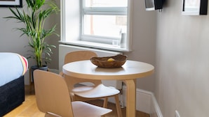 Une petite pièce avec une table ronde en bois, deux chaises, un lit, une plante en pot et une fenêtre donnant sur un paysage agréable.