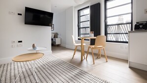 A modern, well-lit living room with a flat-screen TV, a dining area with a table and chairs, and large windows offering a view of the cityscape. - Calm Leicester Sq Studio - Netflix & Nespresso (London)