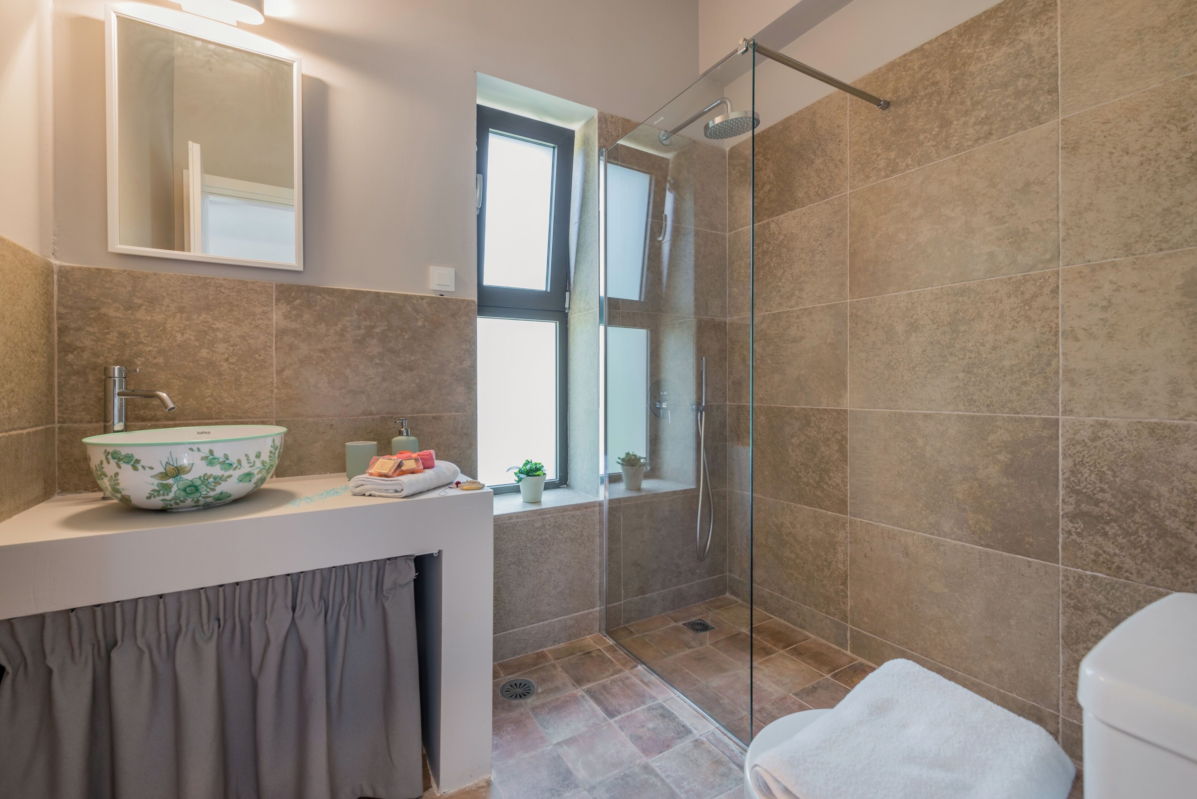 A modern bathroom with a glass shower enclosure, a large mirror, and a window providing natural light.