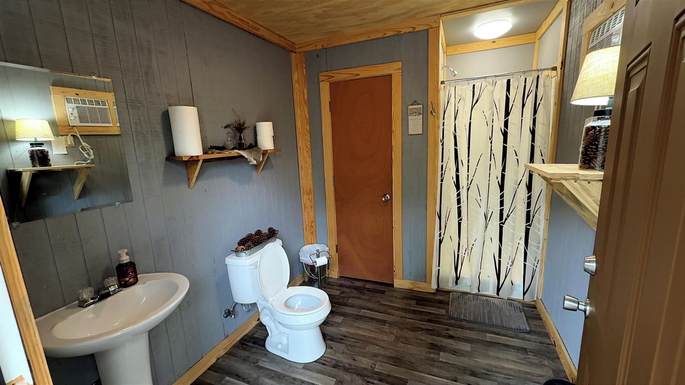A bathroom with a wooden floor, a white sink, a toilet, and a shower curtain with a tree pattern.
