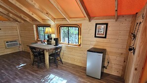A cozy cabin with a wooden interior, a dining area with a table and chairs, a small refrigerator, and a window with a view of trees. - Ozarks Adventure Lodging, Glamping (Ozone)