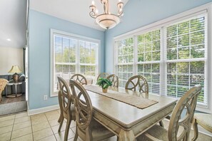 A dining room with a large table, chairs, a chandelier, and large windows with blinds.