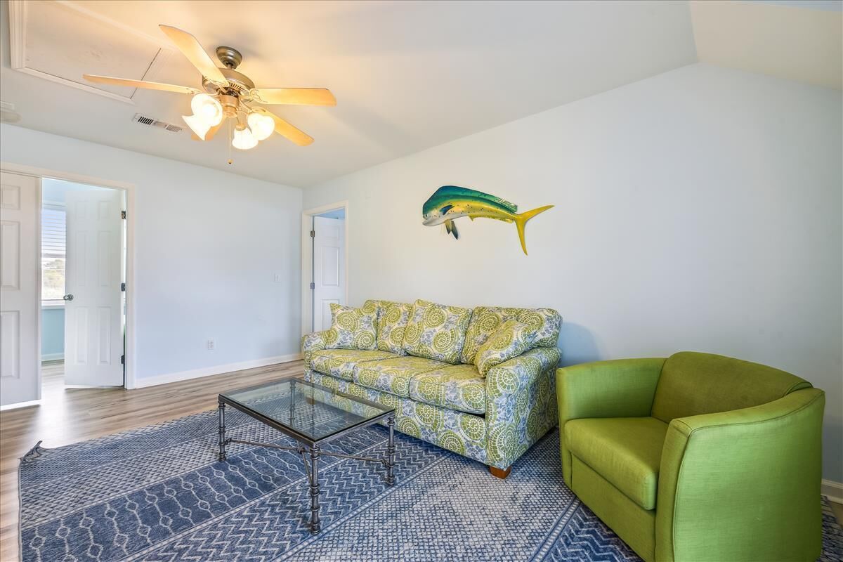 Un soggiorno con un divano a fantasia, una poltrona verde, un tavolino in vetro, una ventola a soffitto e una decorazione a forma di pesce appesa al muro.