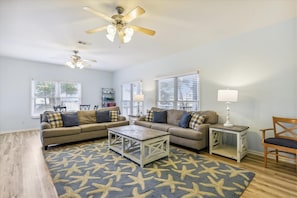 A living room with two sofas, a coffee table, and a patterned rug. There are ceiling fans, windows with blinds, and a lamp on a side table. - A BLUE HORIZON, A perfect getaway anytime of the year! (St. George Island)