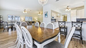 Un comedor amplio con una mesa de madera y sillas blancas, una araña y vistas a la cocina y la sala de estar.