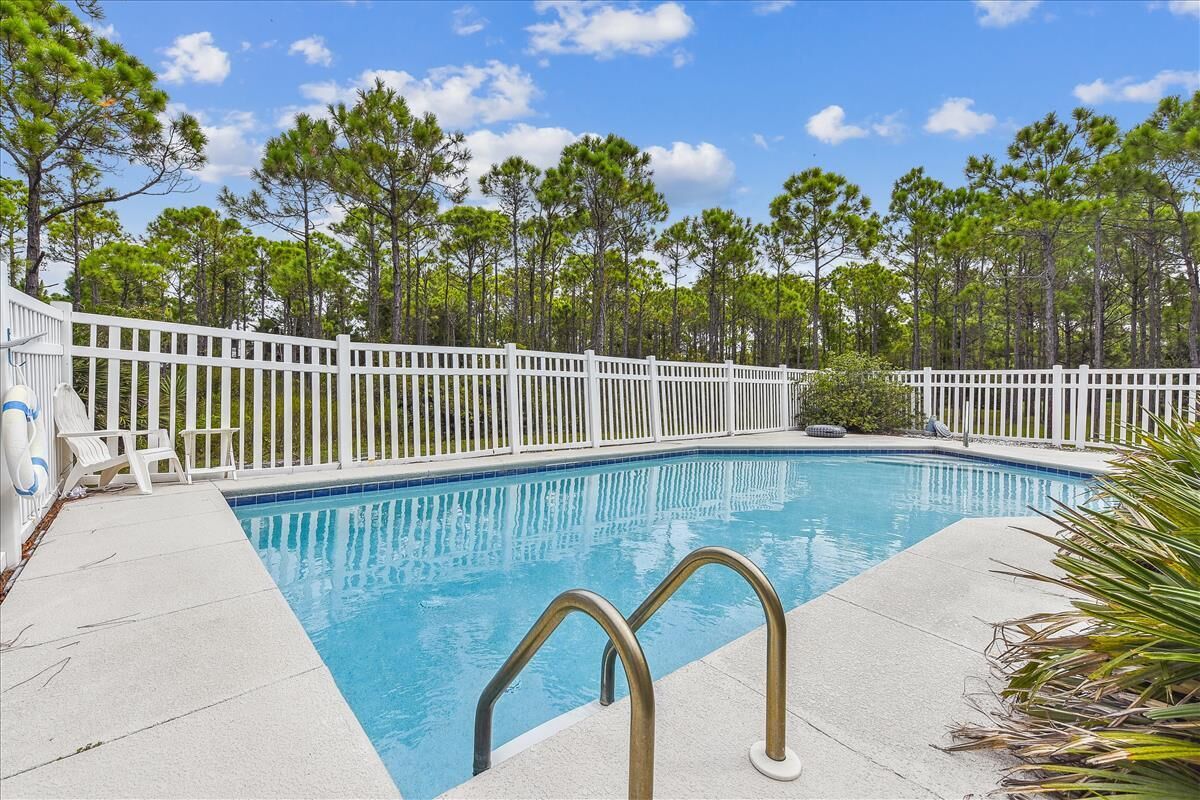 A rectangular swimming pool with a white fence and metal handrails, surrounded by a concrete deck and white lounge chairs.