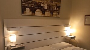 A hotel room with a large bed, two bedside lamps, a small table with a remote control, and a painting of a cityscape on the wall.