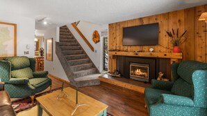 A living room with a fireplace, green patterned chairs, a wooden staircase, and a flat-screen TV mounted on the wall. - Homestead 1: Cozy 2BR in West Yellowstone, just 1 minute from Yellowstone Park! (West Yellowstone)