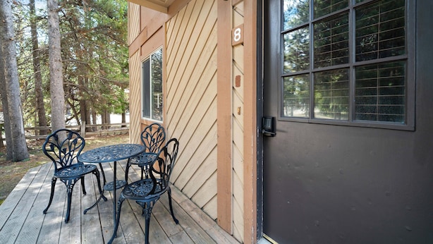 A wooden deck with a table and chairs, a door with a number, and a window with a view of trees.