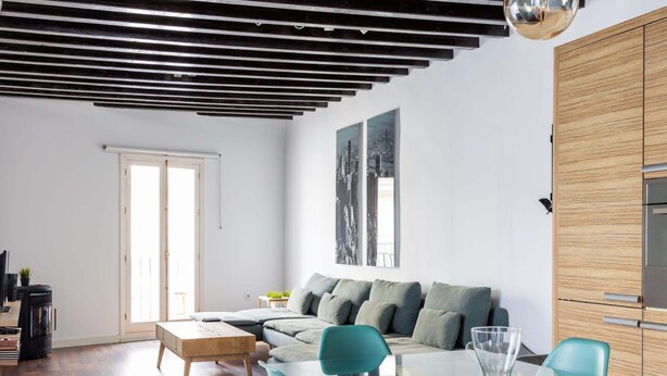 Un soggiorno moderno con un tavolo da pranzo in vetro, sedie color petrolio, un tavolino in legno e un divano con cuscini grigi.