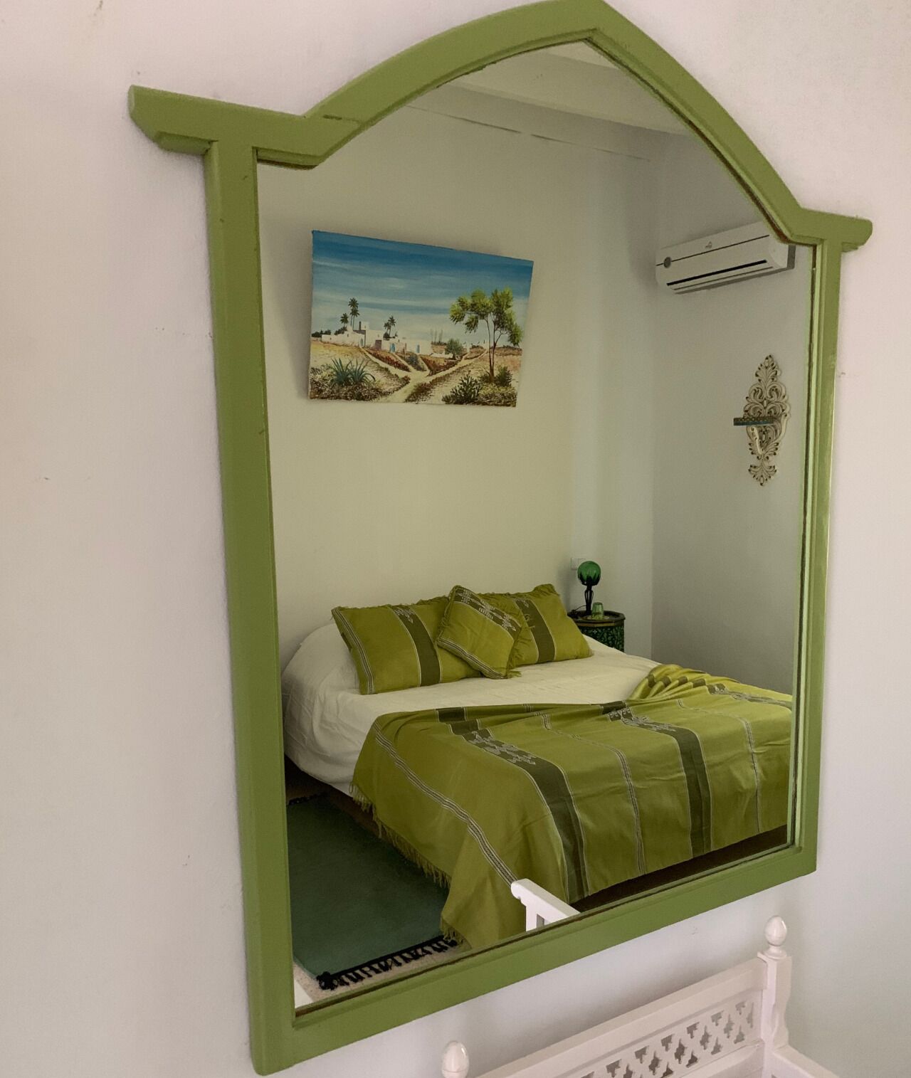 A bedroom with a bed, green bedding, a nightstand, a lamp, a wall-mounted air conditioner, and a painting of a landscape.
