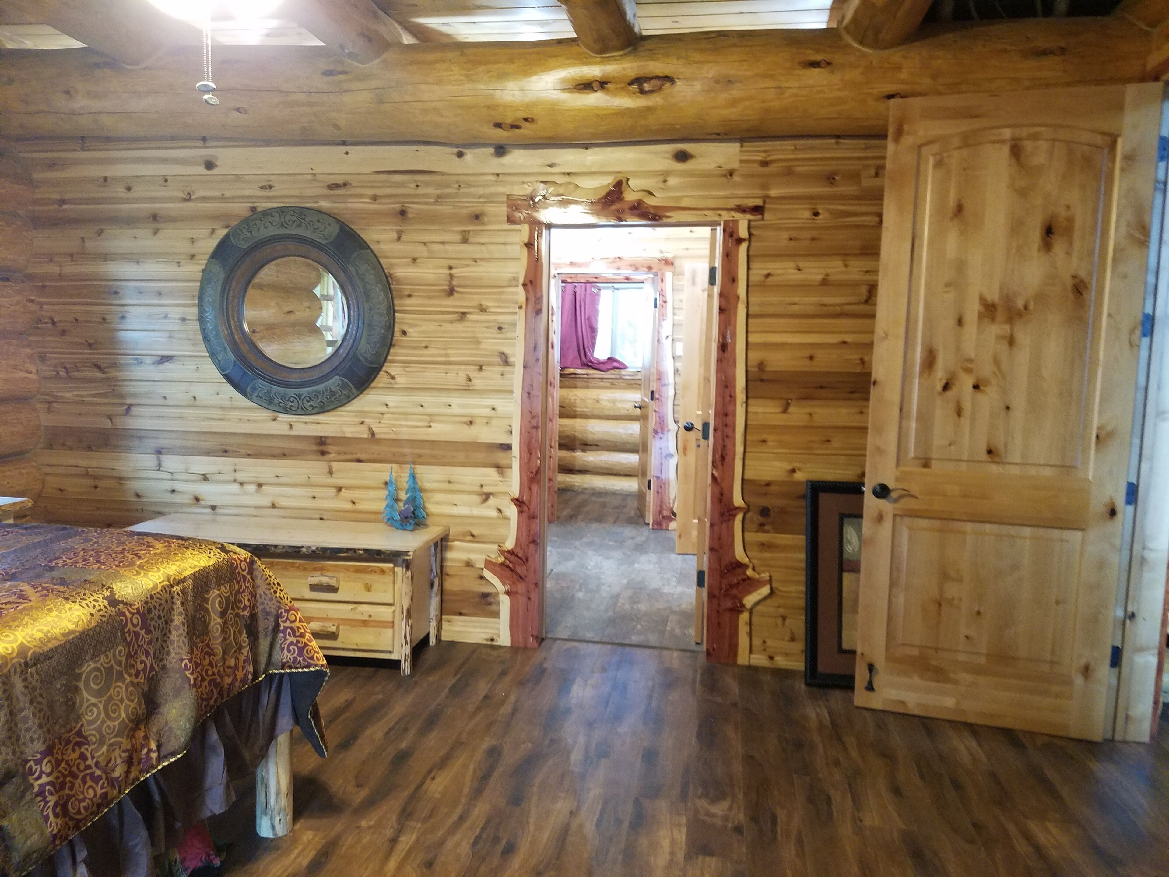 Una habitación tipo cabaña de madera con una cama, un aparador, un espejo y una puerta que da a otra habitación.