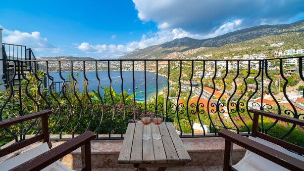 A balcony with a table and two wine glasses, overlooking a coastal town and the sea.