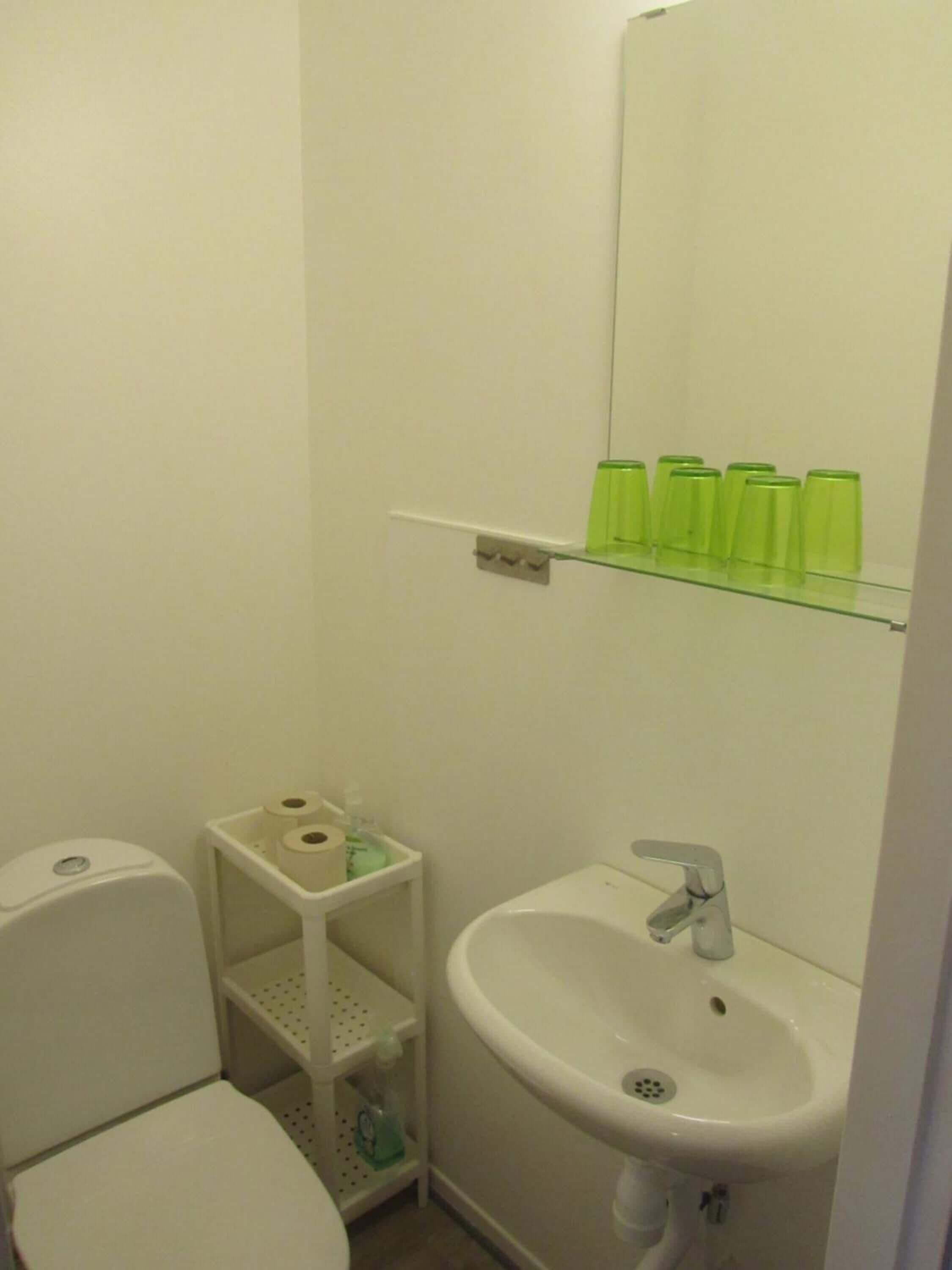 Un baño pequeño con inodoro, lavamanos y espejo. Hay vasos verdes en un estante sobre el lavamanos.