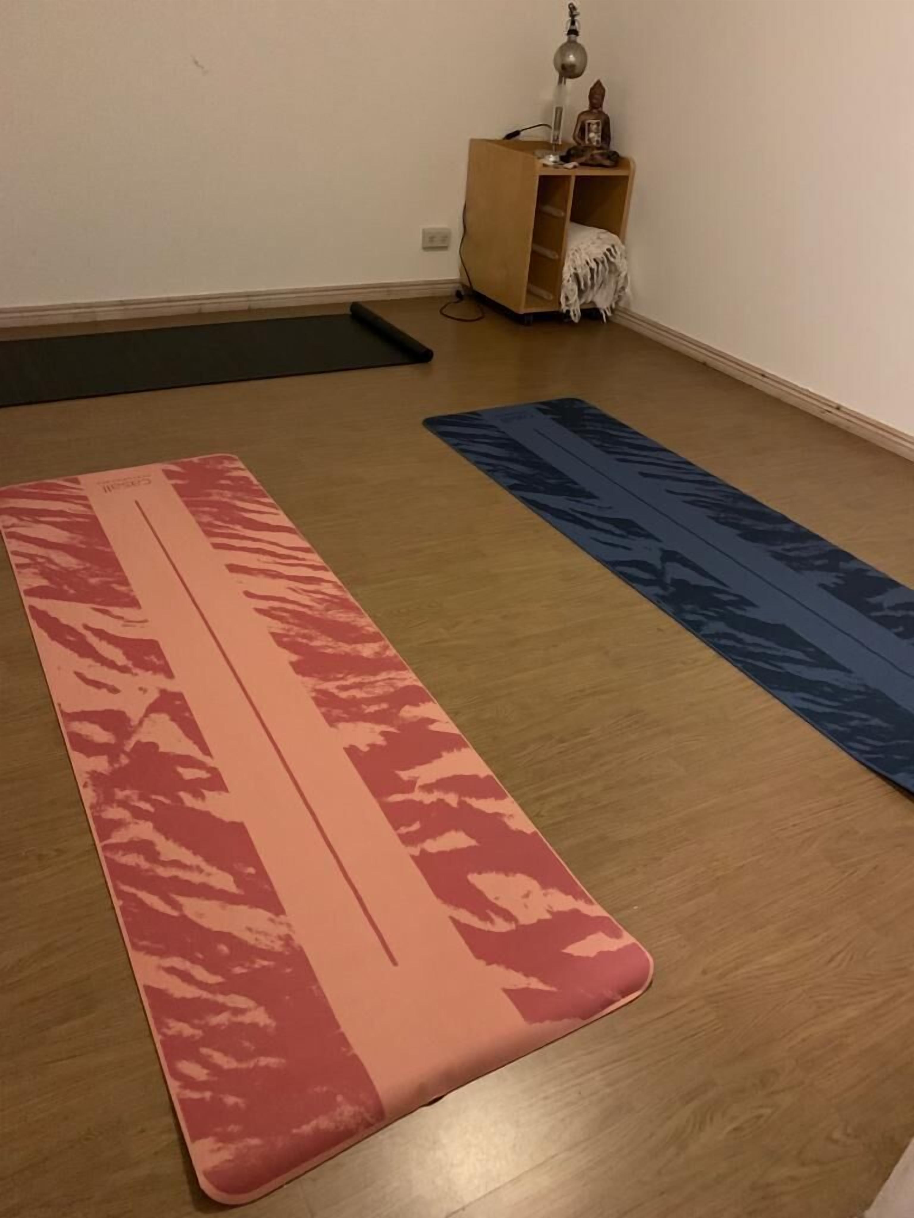Una habitación con piso de madera, una colchoneta de yoga con estampado rosa y rojo, otra colchoneta de yoga con estampado azul y un pequeño mueble de madera con una lámpara y objetos decorativos.