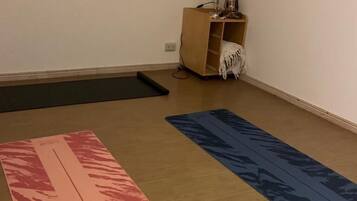 Una habitación con piso de madera, una colchoneta de yoga con estampado rosa y rojo, otra colchoneta de yoga con estampado azul y un pequeño mueble de madera con una lámpara y objetos decorativos.
