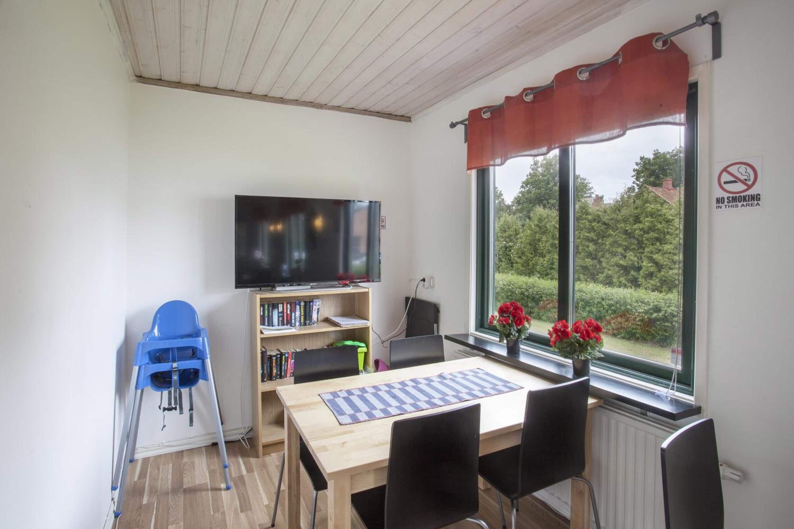 Una habitación con una mesa de madera, sillas negras, una trona azul, una estantería, un televisor y una ventana con cortina roja.
