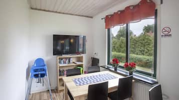 Una habitación con una mesa de madera, sillas negras, una trona azul, una estantería, un televisor y una ventana con cortina roja.