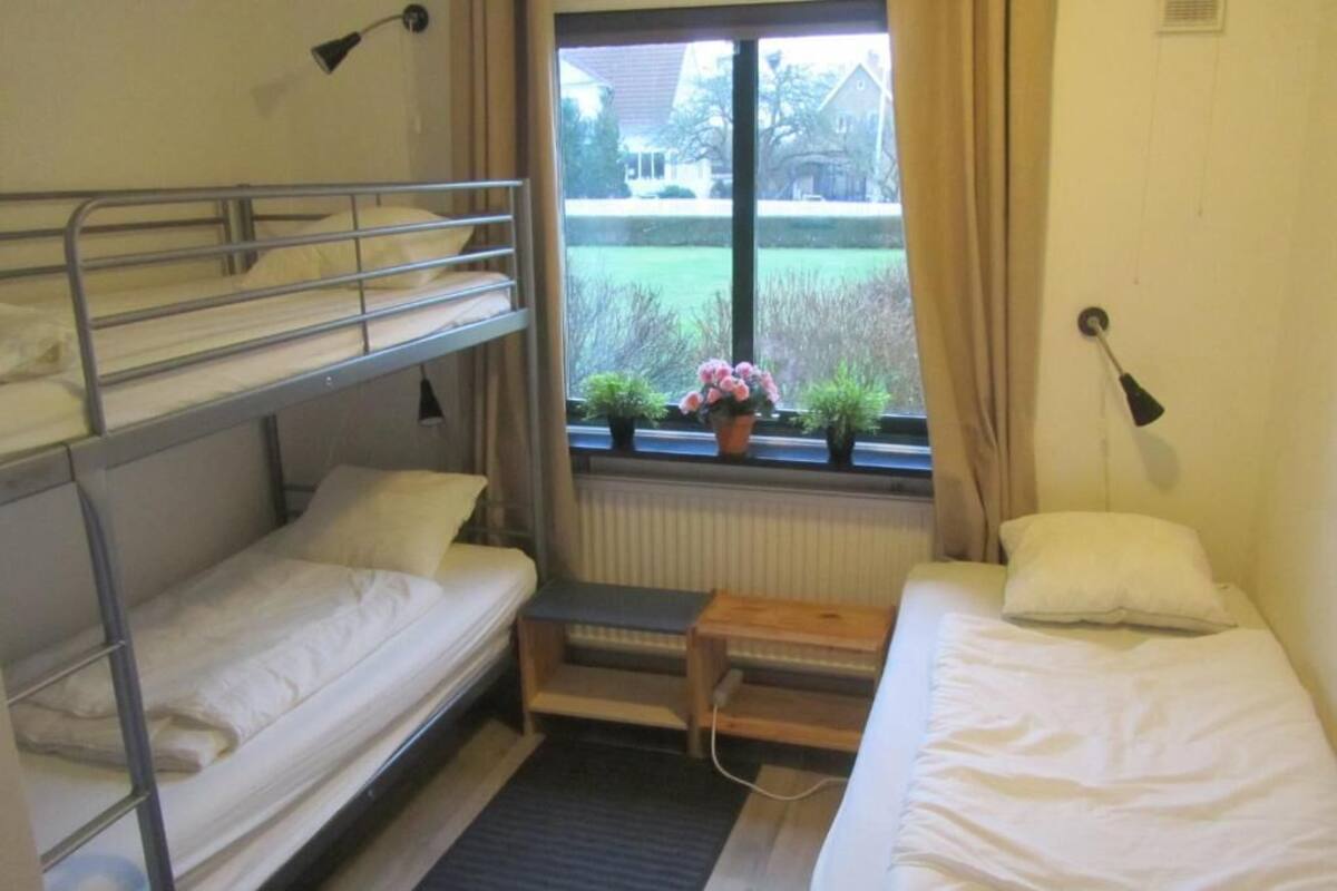 Um dormitório com beliches, uma mesinha, uma janela com cortinas e vista para o exterior.