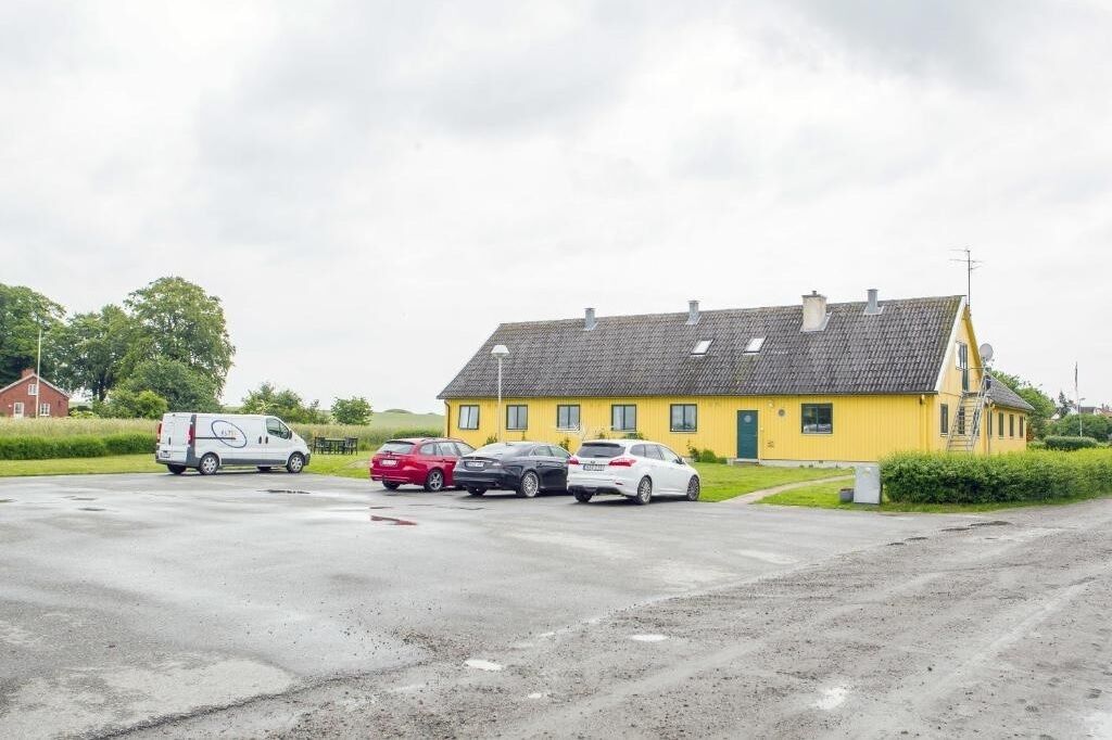 Un bâtiment jaune avec un toit en pente, un fourgon blanc et plusieurs voitures garées devant.