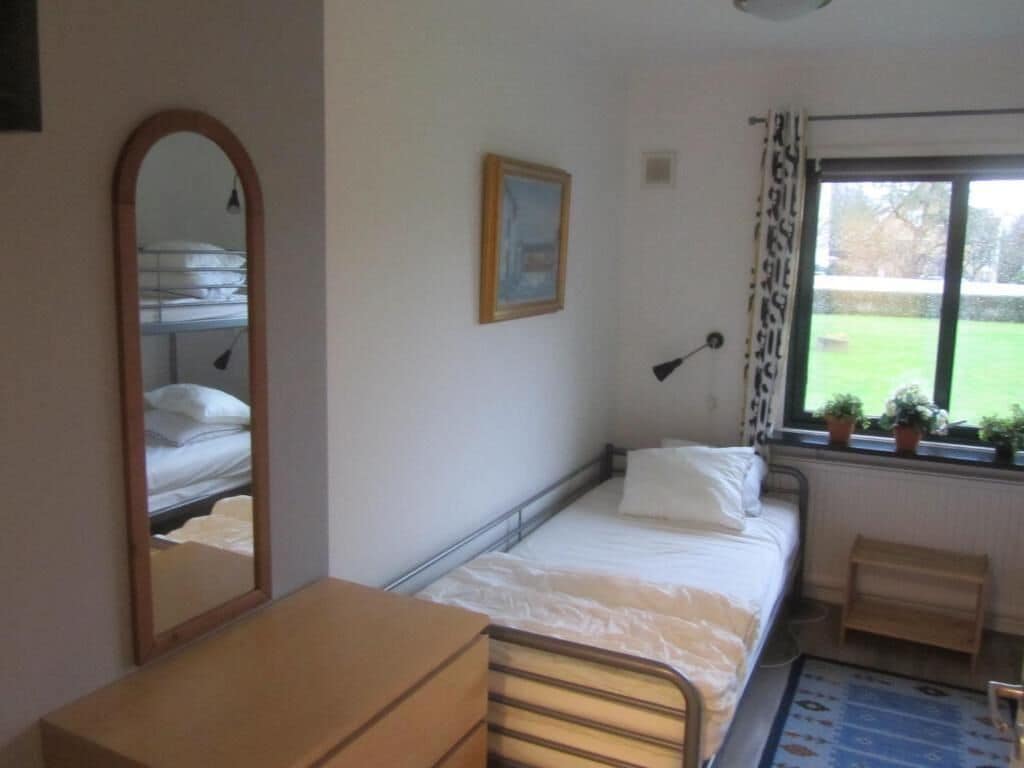 A bedroom with a single bed, a bedside table, a mirror, a window with curtains, and a radiator.