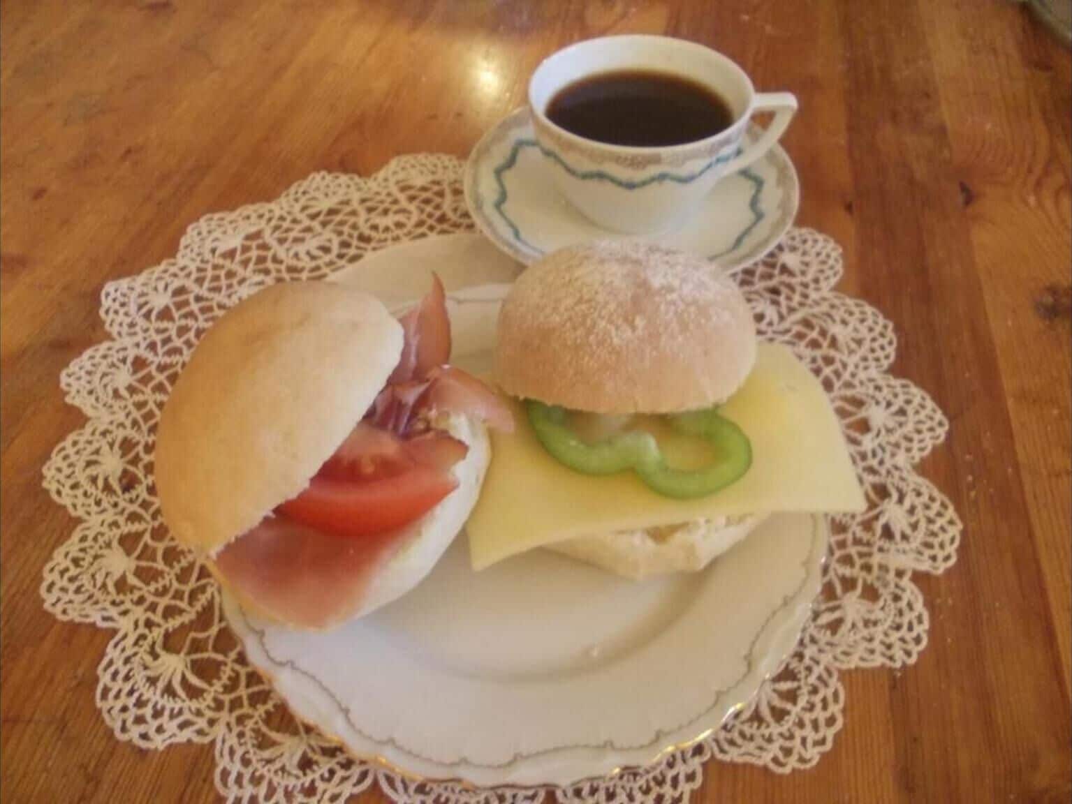 Un sandwich au jambon, à la tomate et au fromage, servi dans un pain, une tasse de café et une assiette blanche ornée d’une serviette en dentelle.