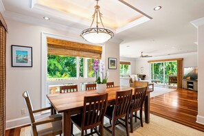 A spacious dining area with a wooden table and chairs, large windows with views of greenery, and a ceiling fan. - Hanalei Ohana, The Ultimate Hanalei Home TVNC#5128 (Hanalei)