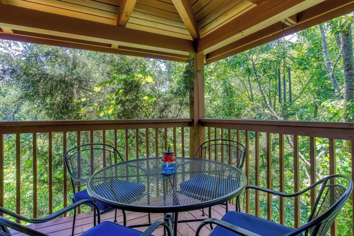 A covered outdoor area with a round metal table and four chairs, surrounded by a wooden railing, overlooking a dense forest.