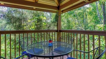 A covered outdoor area with a round metal table and four chairs, surrounded by a wooden railing, overlooking a dense forest.