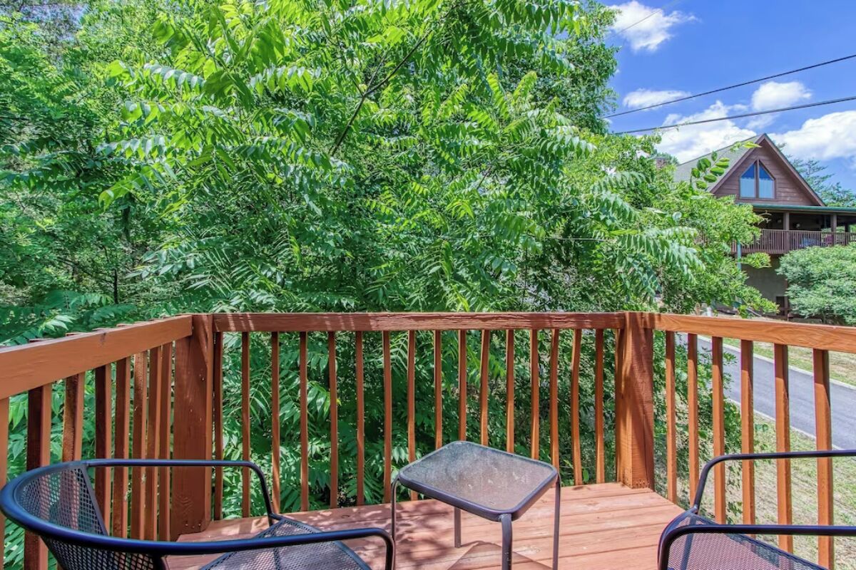 Una terraza de madera con dos sillas de metal y una pequeña mesa, rodeada de abundante follaje verde.