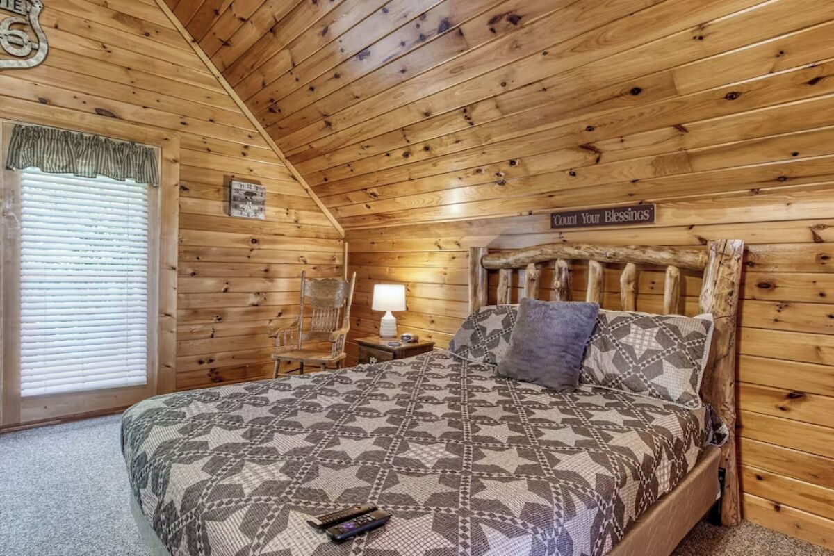 Camera accogliente in legno con un letto, un comodino, una lampada e un cartello che recita 'Comfort Your Blessings'.