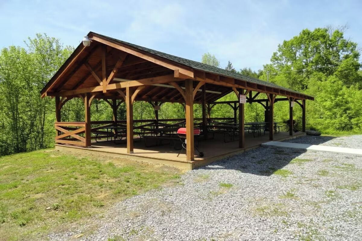 A wooden pavilion with picnic tables and a barbecue grill, surrounded by gravel and greenery.