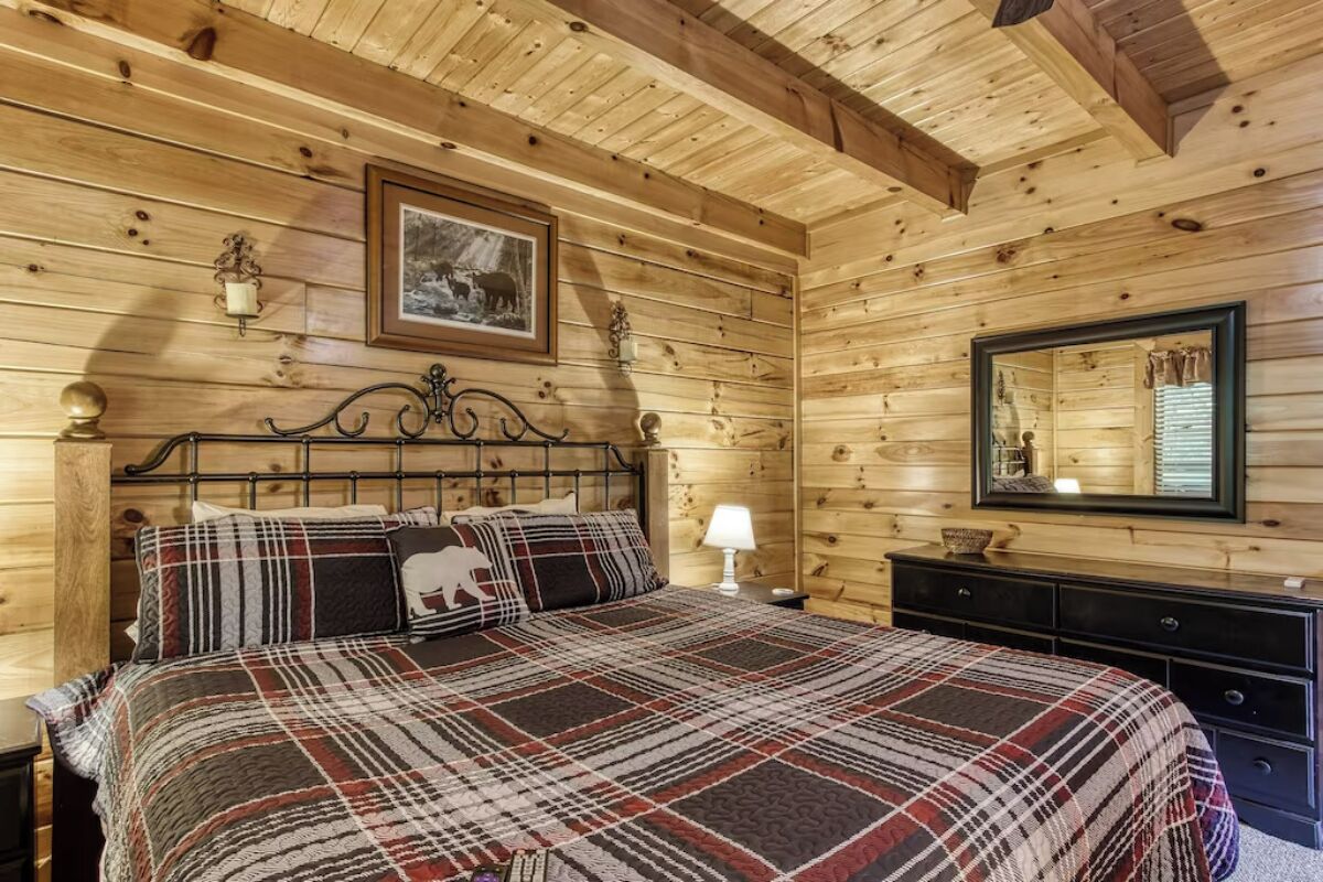 A wooden cabin bedroom with a bed, a nightstand, a dresser, a mirror, and a framed picture on the wall.