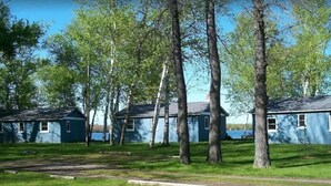 A row of blue cabins with a grassy lawn and trees in the foreground. - Presque Isle Resort, cottage #3 of 3 (Alpena)