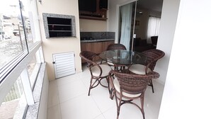 A dining area with wicker chairs and a glass table, a kitchen with granite countertops, and a sliding glass door leading to a balcony. - 315, Ótimo Apartamento 3 Dormitórios Próximo a Praia (SC)