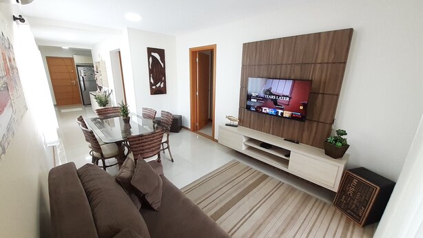 A modern living room with a brown sofa, a glass dining table, and a TV mounted on a wooden panel wall. - 315, Ótimo Apartamento 3 Dormitórios Próximo a Praia (SC)