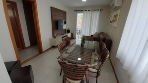 A dining area with a glass-top table and wicker chairs, a TV mounted on the wall, and a balcony with curtains. - 315, Ótimo Apartamento 3 Dormitórios Próximo a Praia (SC)