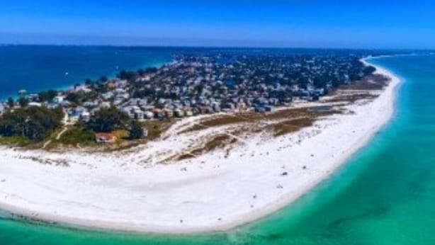 Ett flygbildsbild av ett kustområde med en lång strand, ett tättbebyggt bostadsområde och en klarblå himmel.