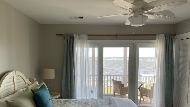 A bedroom with a bed, a ceiling fan, a view of the ocean through a sliding glass door, and a balcony with a table and chairs.