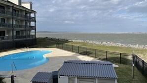 A pool area with a view of the ocean, a building with balconies, and a fenced beach area.