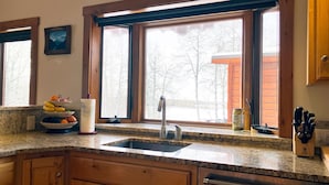 Una cocina con encimeras de granito, gabinetes de madera, un lavamanos de acero inoxidable y una ventana con vistas a árboles y un edificio.