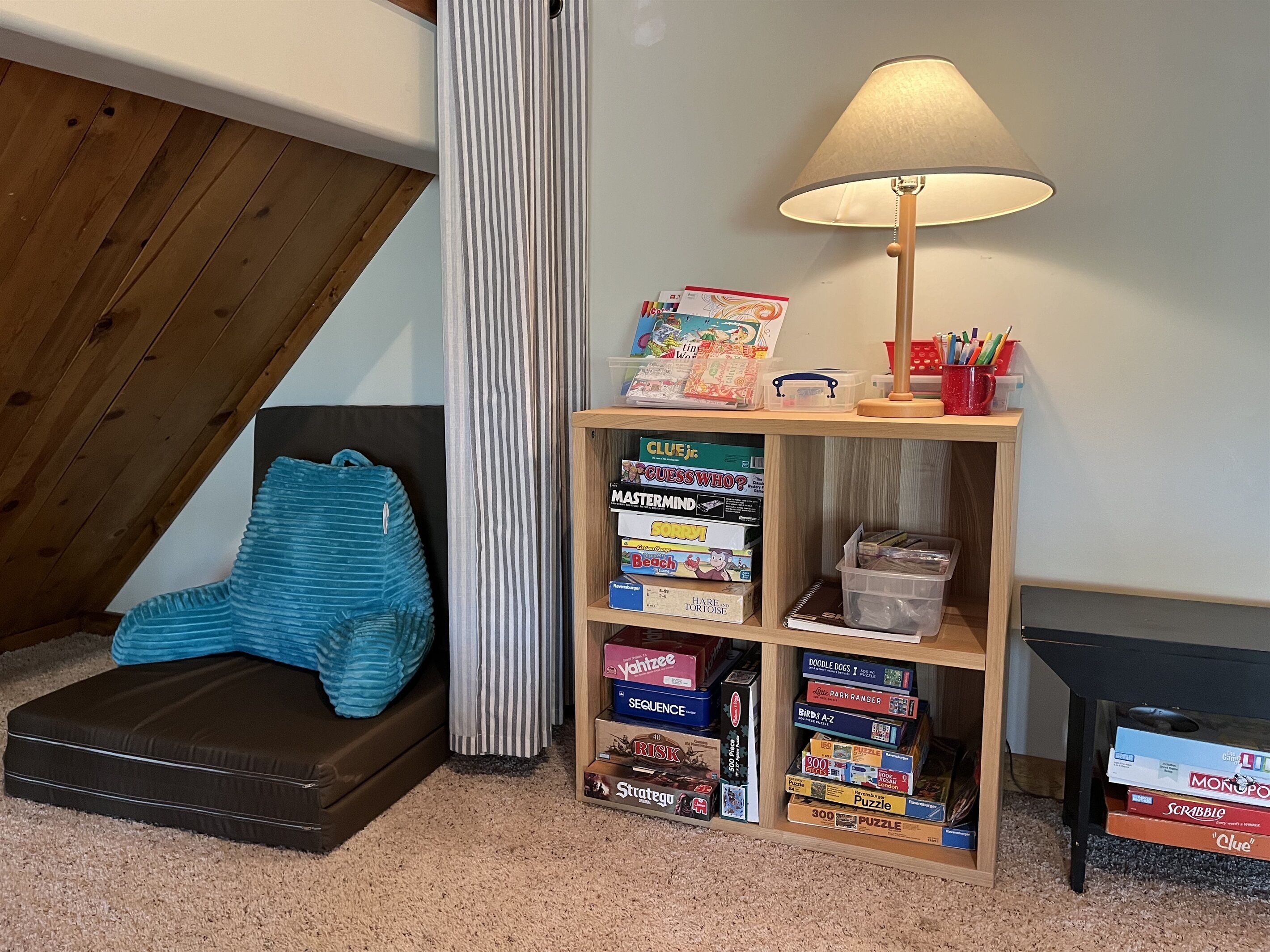 Una habitación acogedora con techo inclinado, una estantería de madera llena de juegos de mesa, una manta azul en un sofá y una lámpara sobre una mesita.