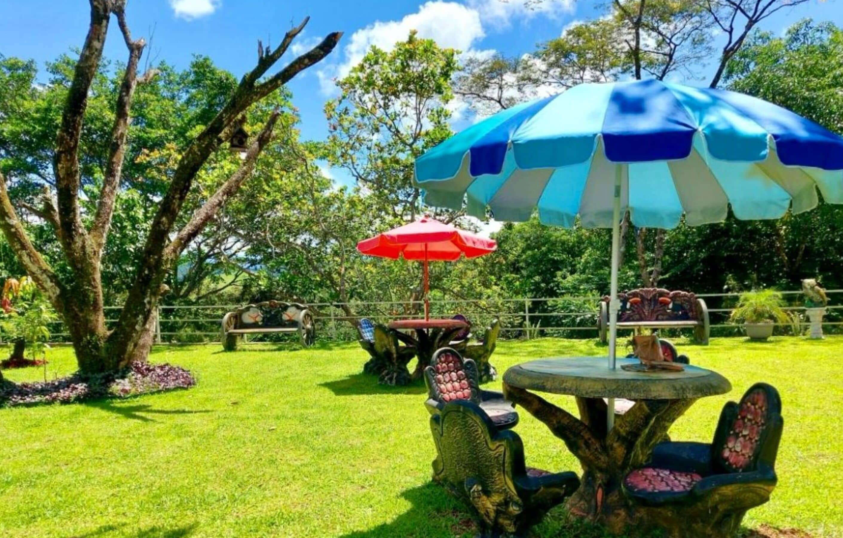 Un jardín con una mesa redonda y sillas, dos sombrillas y sillas de piedra decorativas.