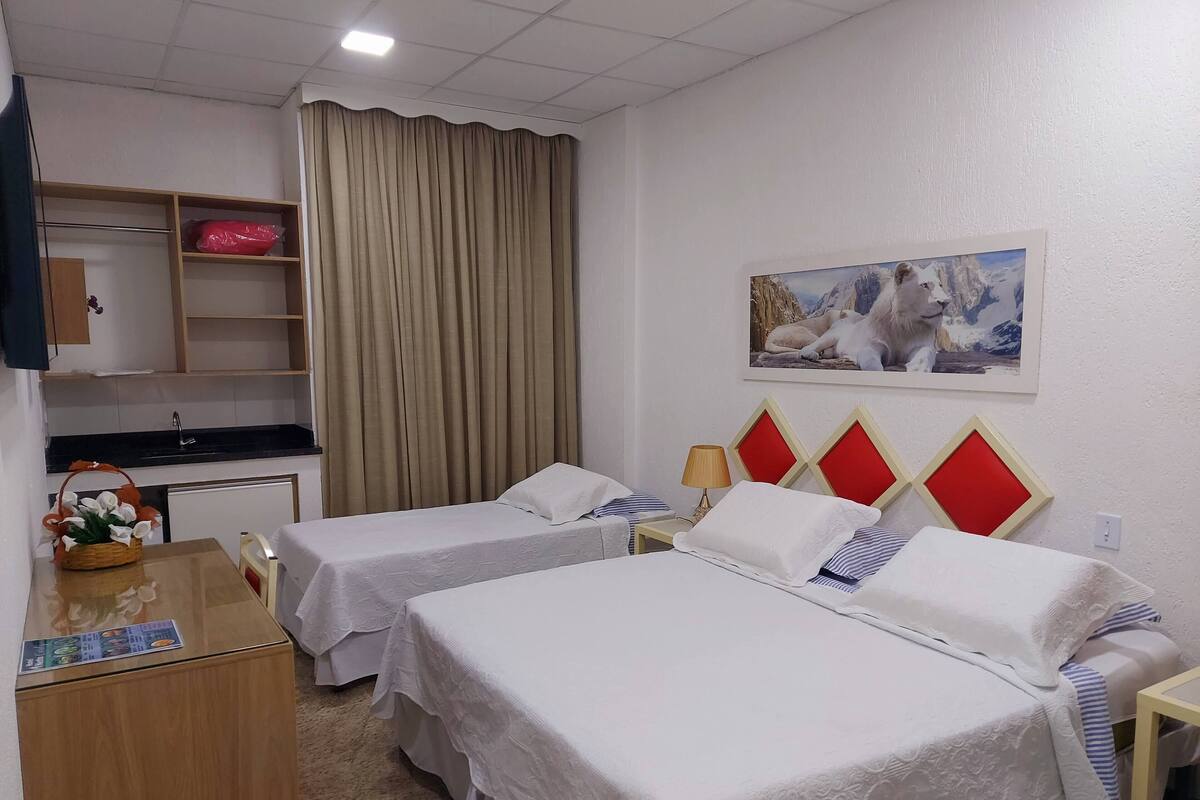 Habitación de hotel con dos camas, un escritorio, una silla, una mesita con una cesta, una cortina y un cuadro en la pared.
