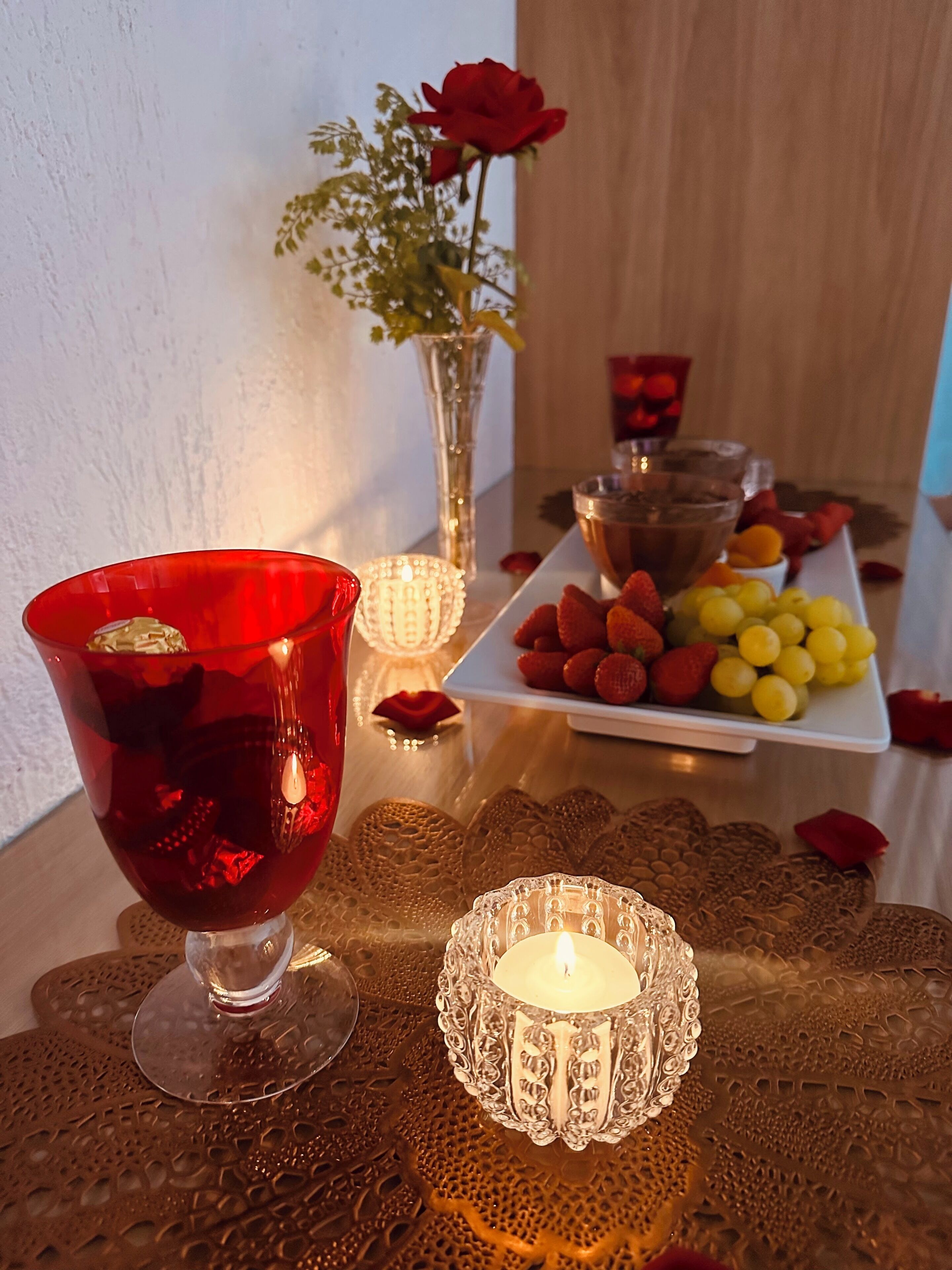 Una mesa con un vaso rojo, una vela encendida, un florero con una rosa roja y un plato de fruta.