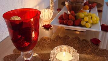 Una mesa con un vaso rojo, una vela encendida, un florero con una rosa roja y un plato de fruta.